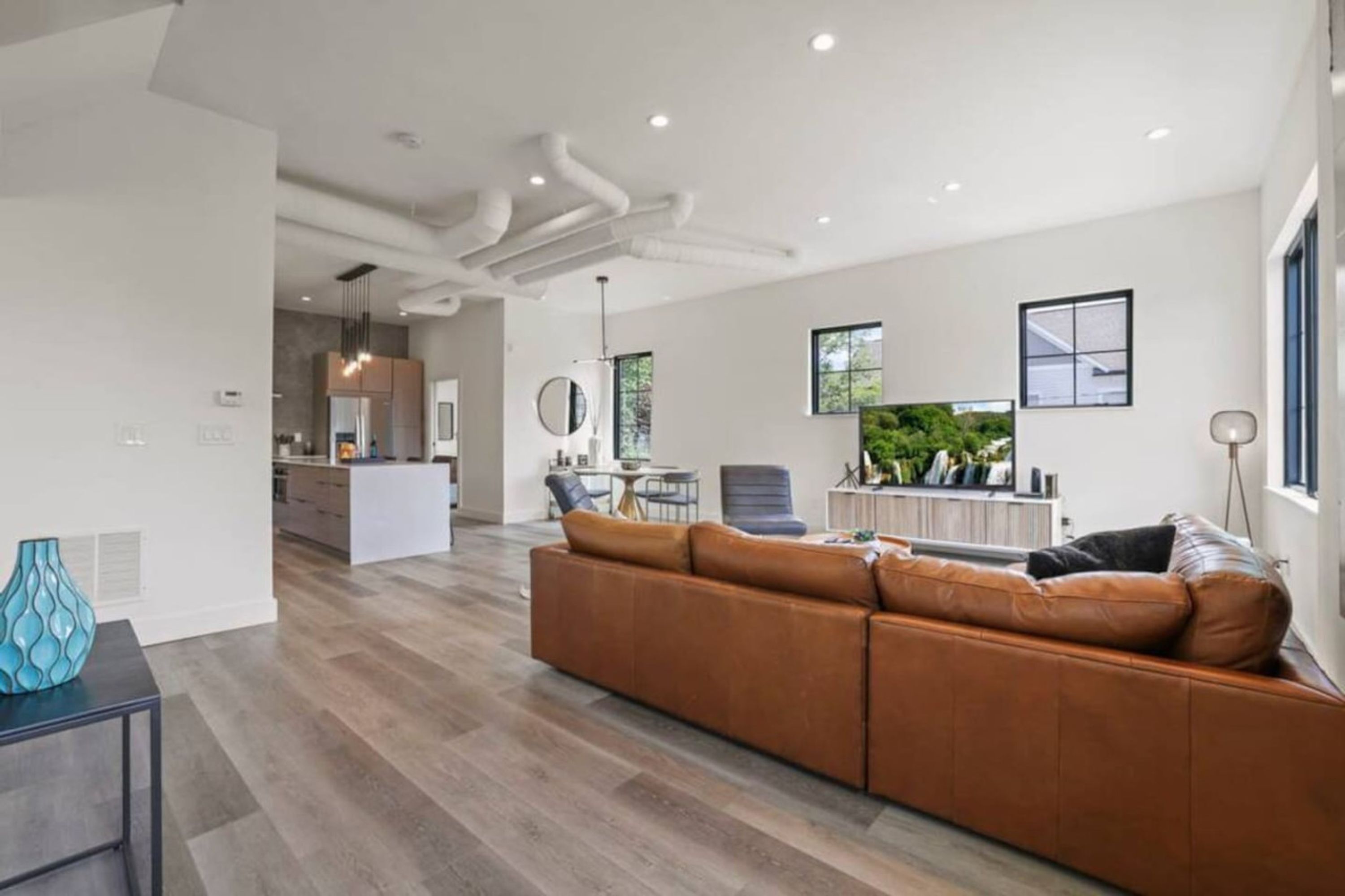 A modern living room with a brown leather sofa, a dining area with a round table, and a kitchen in the background.