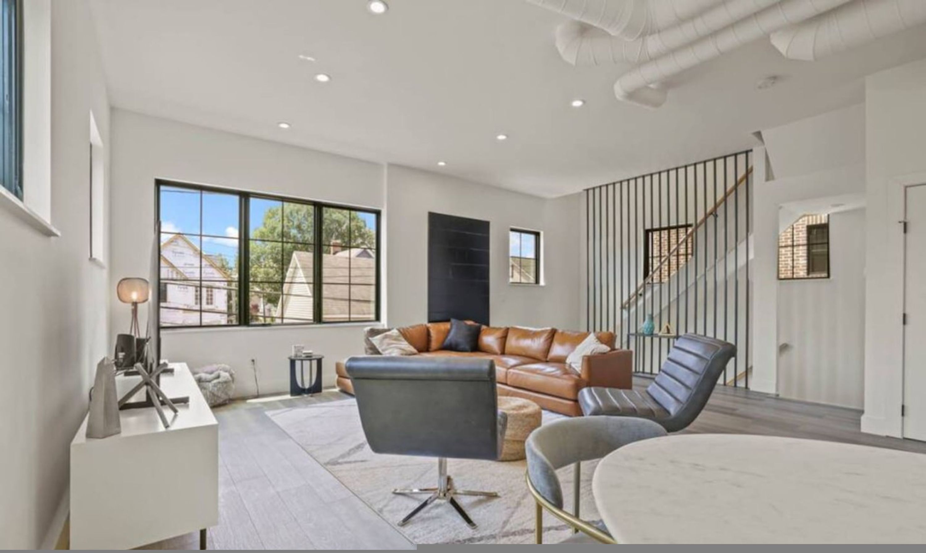 A spacious living room with a leather sofa, a coffee table, and a modern chair. There is a staircase with a railing and a view of the outside through large windows.