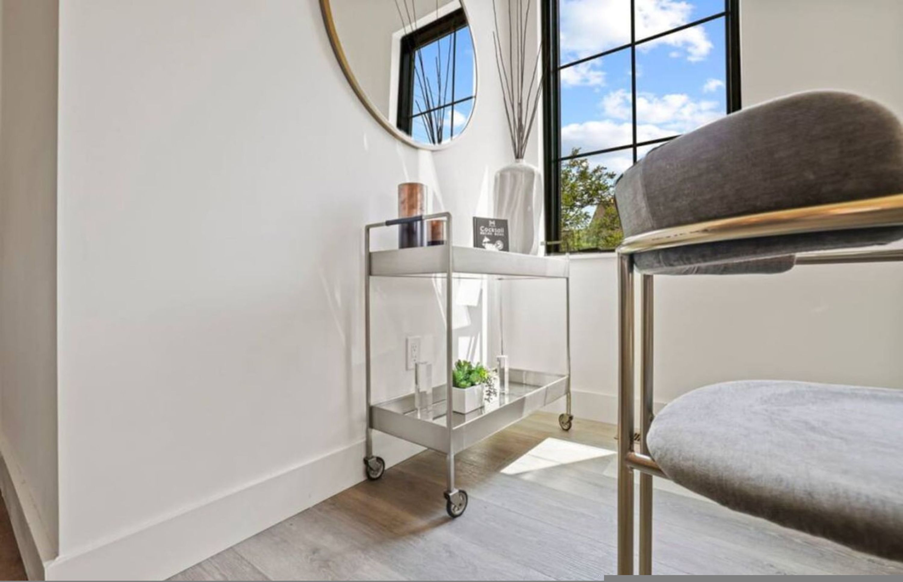 A room with a grey chair, a metal cart with a plant, and a window with a view of trees.