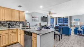 A modern kitchen with wooden cabinets, a dishwasher, and a granite countertop. There is a dining area with blue furniture and a view of the city through large windows. - The Whaler B3 (Gulf Shores)