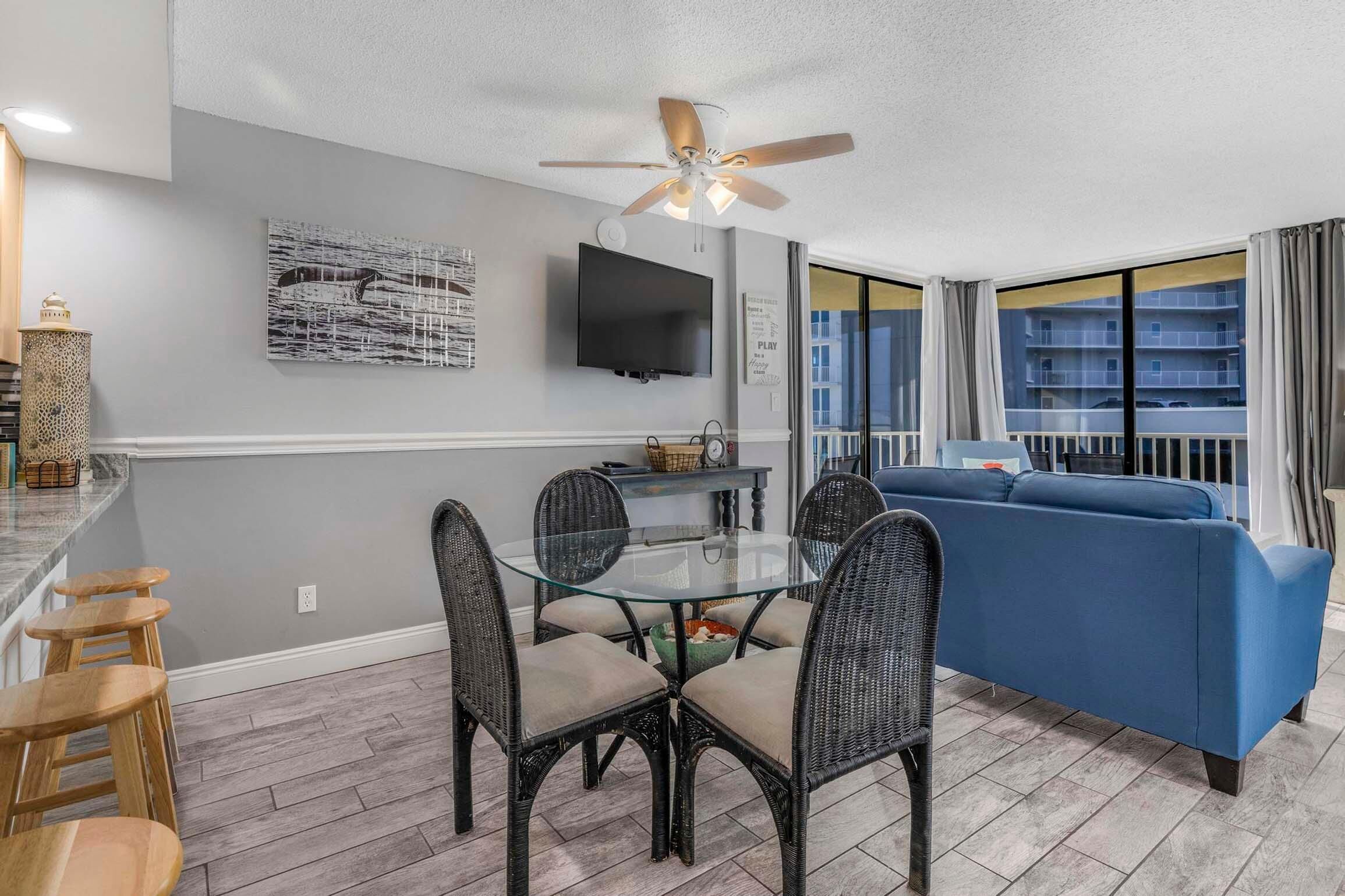 A modern living room with a glass-top dining table, wicker chairs, a blue sofa, and a ceiling fan.
