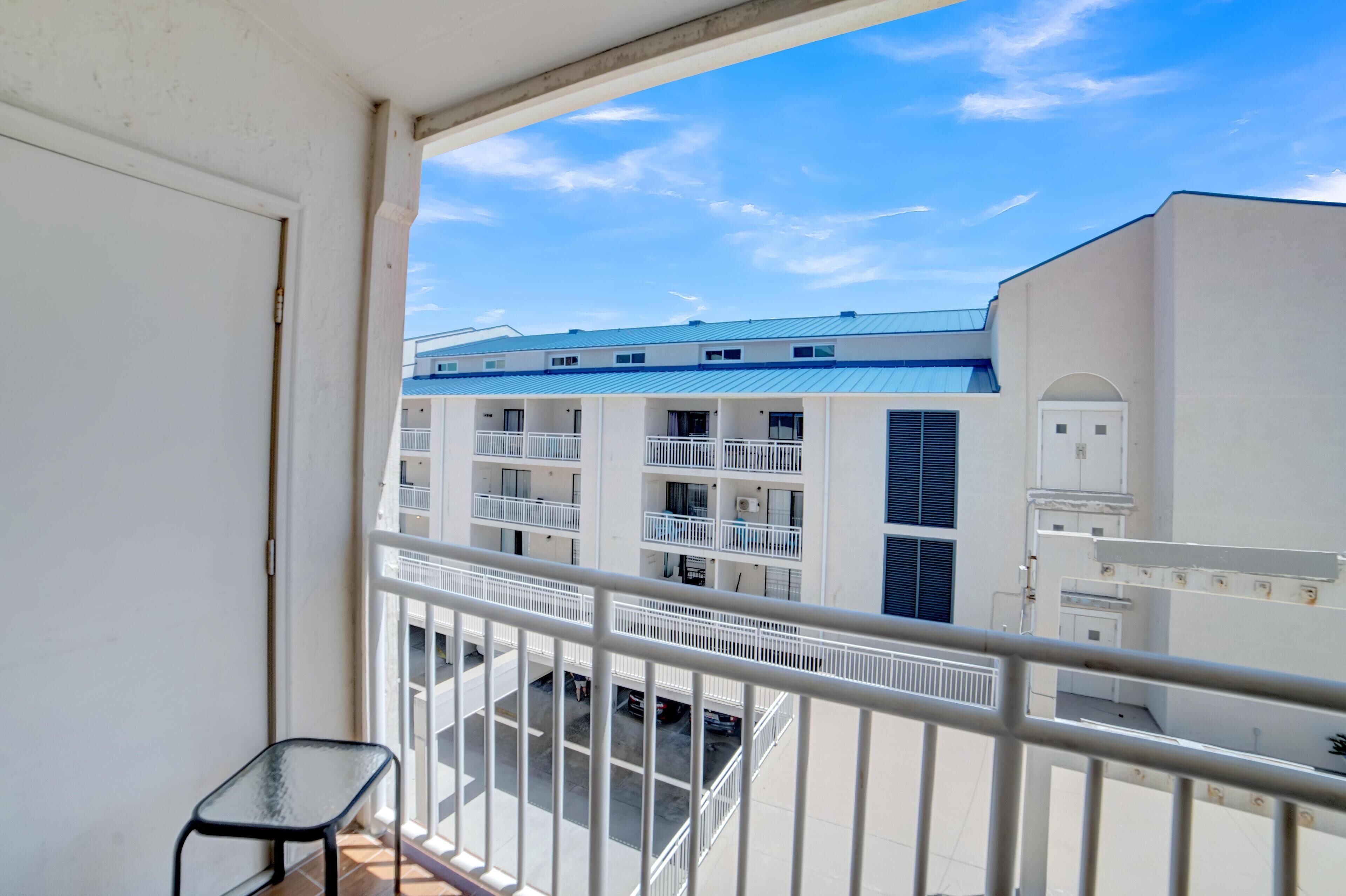 A balcony with a white railing, a small table, and a view of a multi-story building with balconies and a blue roof.
