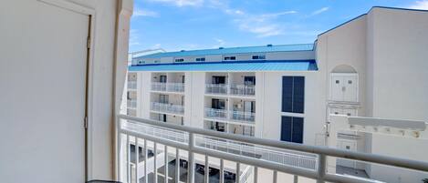A balcony with a white railing, a small table, and a view of a multi-story building with balconies and a blue roof.