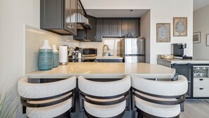 A modern kitchen with grey cabinets, a central island with bar stools, stainless steel appliances, and decorative wall art. - Battleship Vista - Parking (Wilmington)