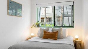 A bedroom with a bed, pillows, a bedside table with a lamp, a window with a view of buildings, and a wall-mounted picture. - Apartment 2 Surfside. Combining an amazing beachside location, just 500m from town (Byron Bay)