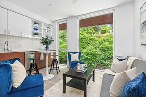 A modern living room with a kitchen, featuring a sofa, a coffee table, and a view of greenery through large windows.