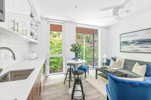 Une cuisine moderne dotée d’une grande fenêtre, un coin repas avec une table ronde et des tabourets, et un salon avec un canapé et un tableau.