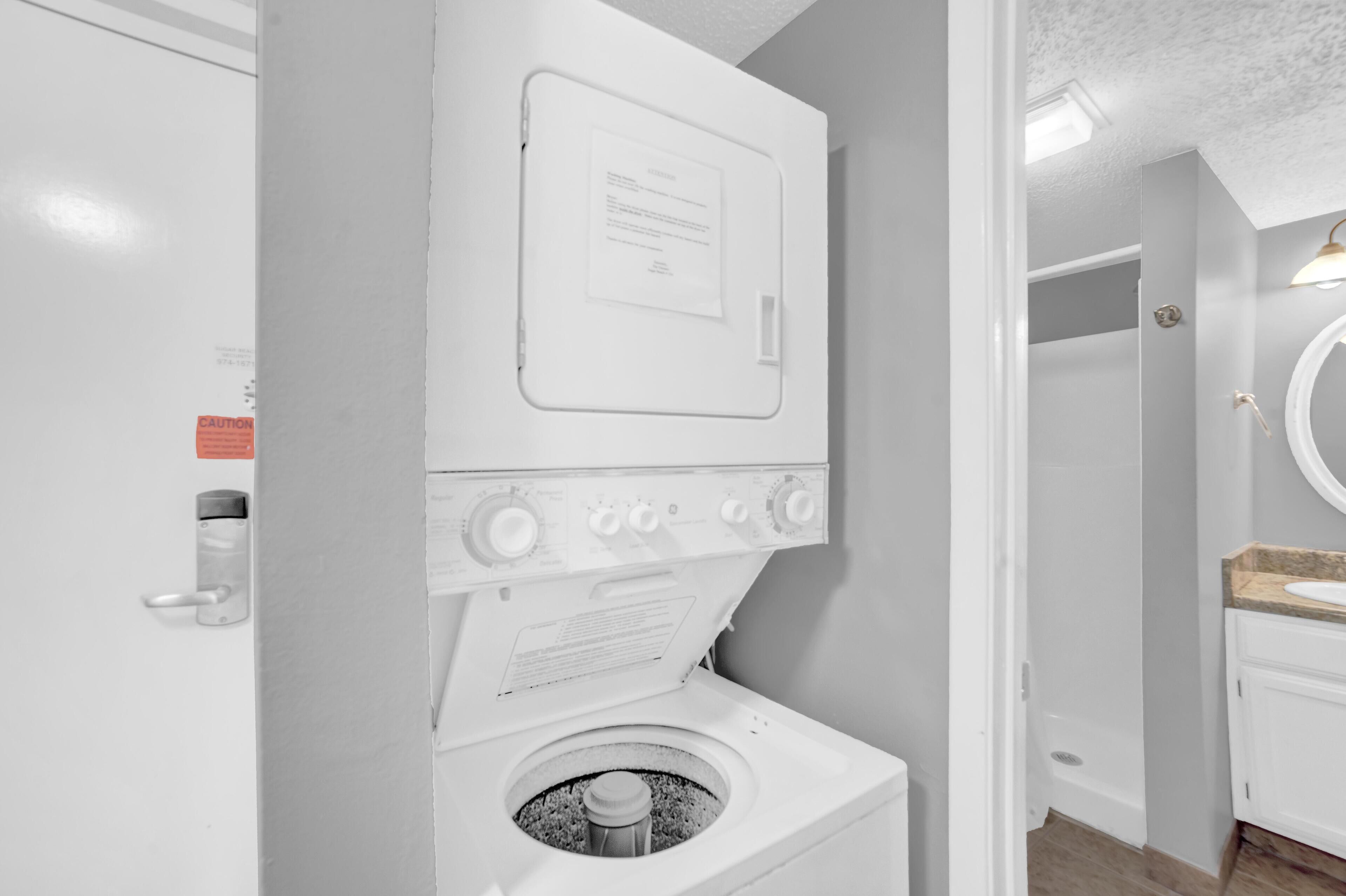 Une petite buanderie équipée d’un lave-linge et d’un sèche-linge à chargement frontal, d’un panneau d’avertissement sur la porte et d’un petit coin lavabo avec un miroir.