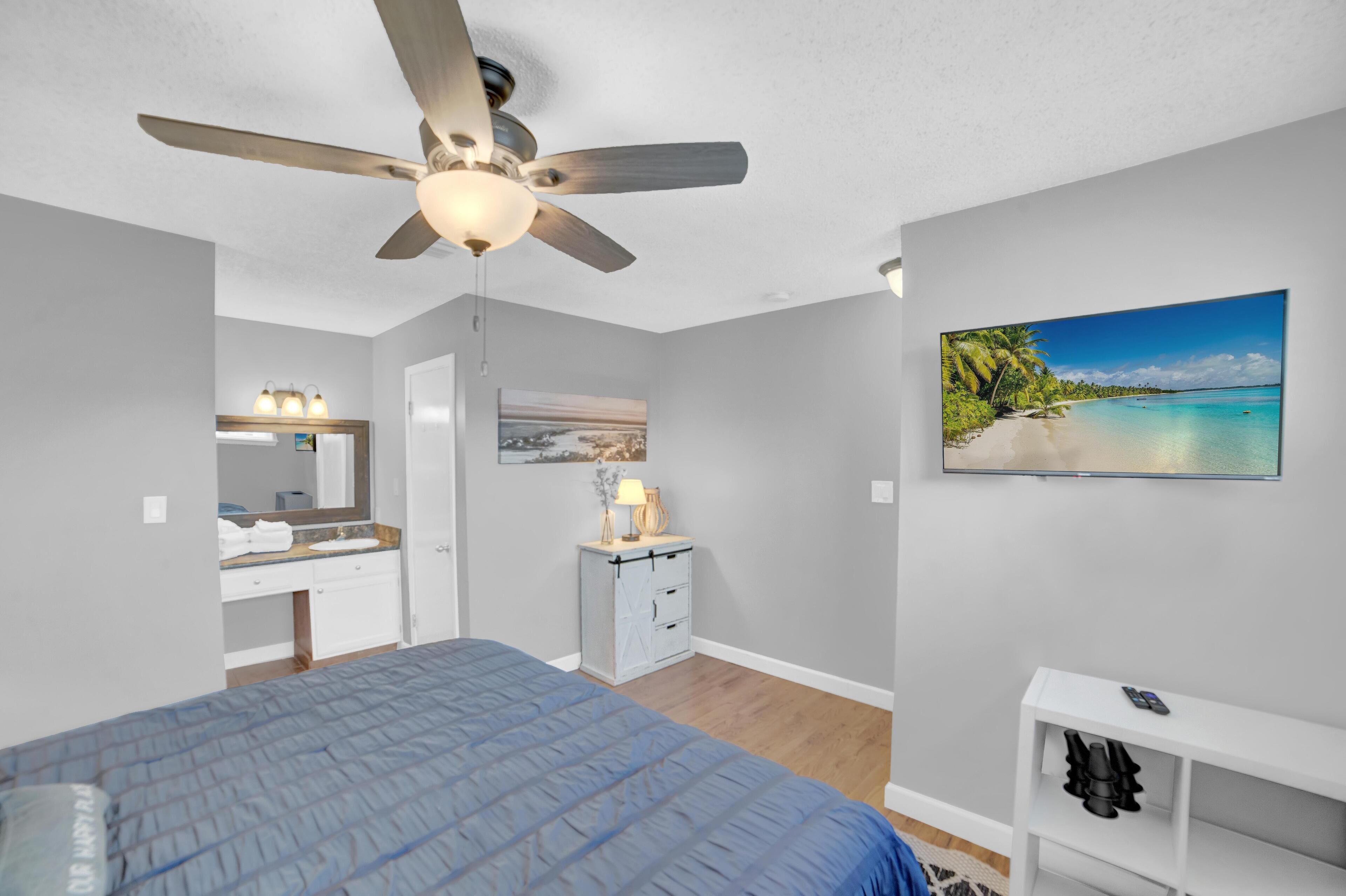 A bedroom with a bed, ceiling fan, bedside table, nightstand, and a painting of a beach scene.