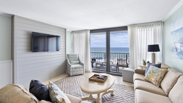A living room with a sofa, armchair, and coffee table, a flat-screen TV mounted on the wall, and a balcony with a view of the ocean. - The Breakers 42 (Orange Beach)