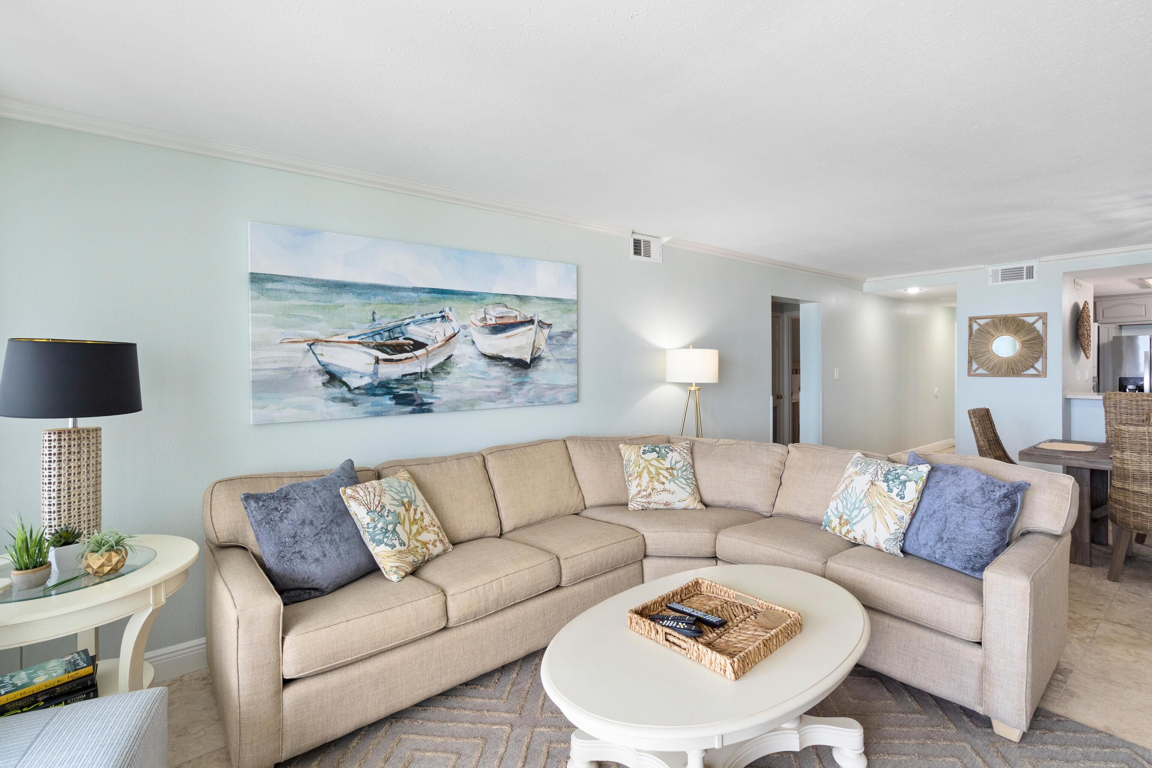 Una sala de estar con un sofá seccional color beige, una mesa redonda, un cuadro con barcos en la pared, una lámpara y una mesita con lámpara.