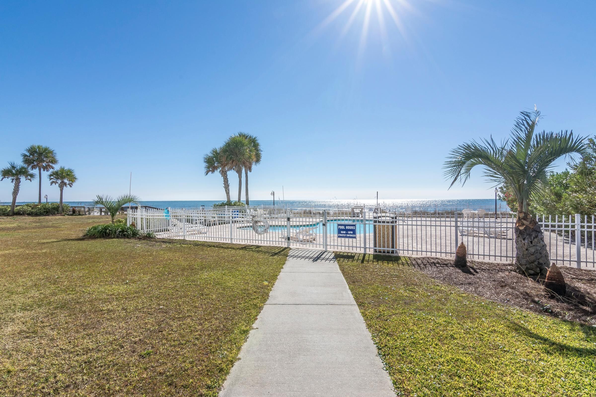 A pathway leading to a fenced pool area with palm trees and a clear view of the ocean.