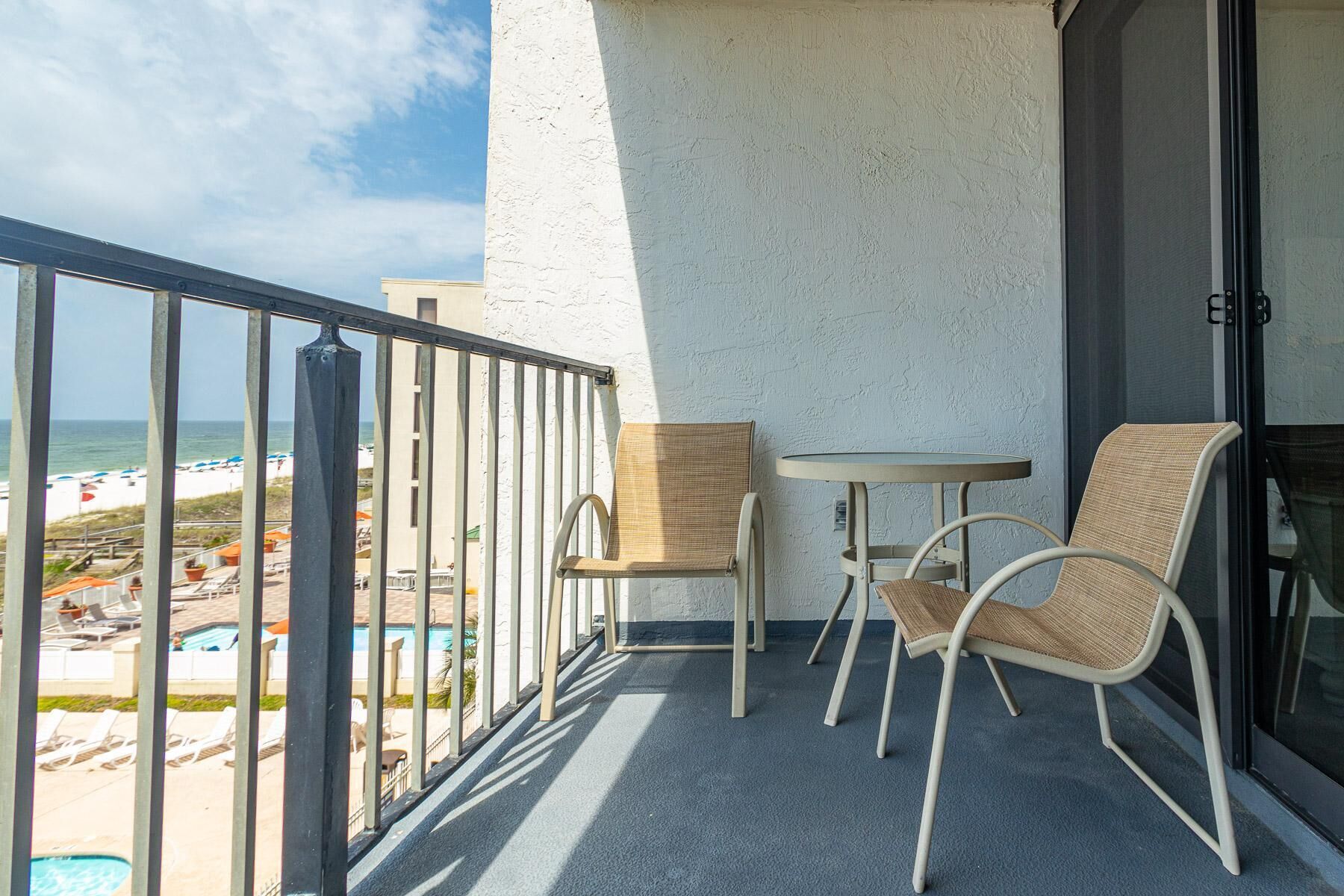 A balcony with two wicker chairs and a small table, overlooking a pool and beach area.
