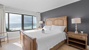 A bedroom with a large window, wooden bed, nightstands, and a view of the beach. - The Breakers 42 (Orange Beach)
