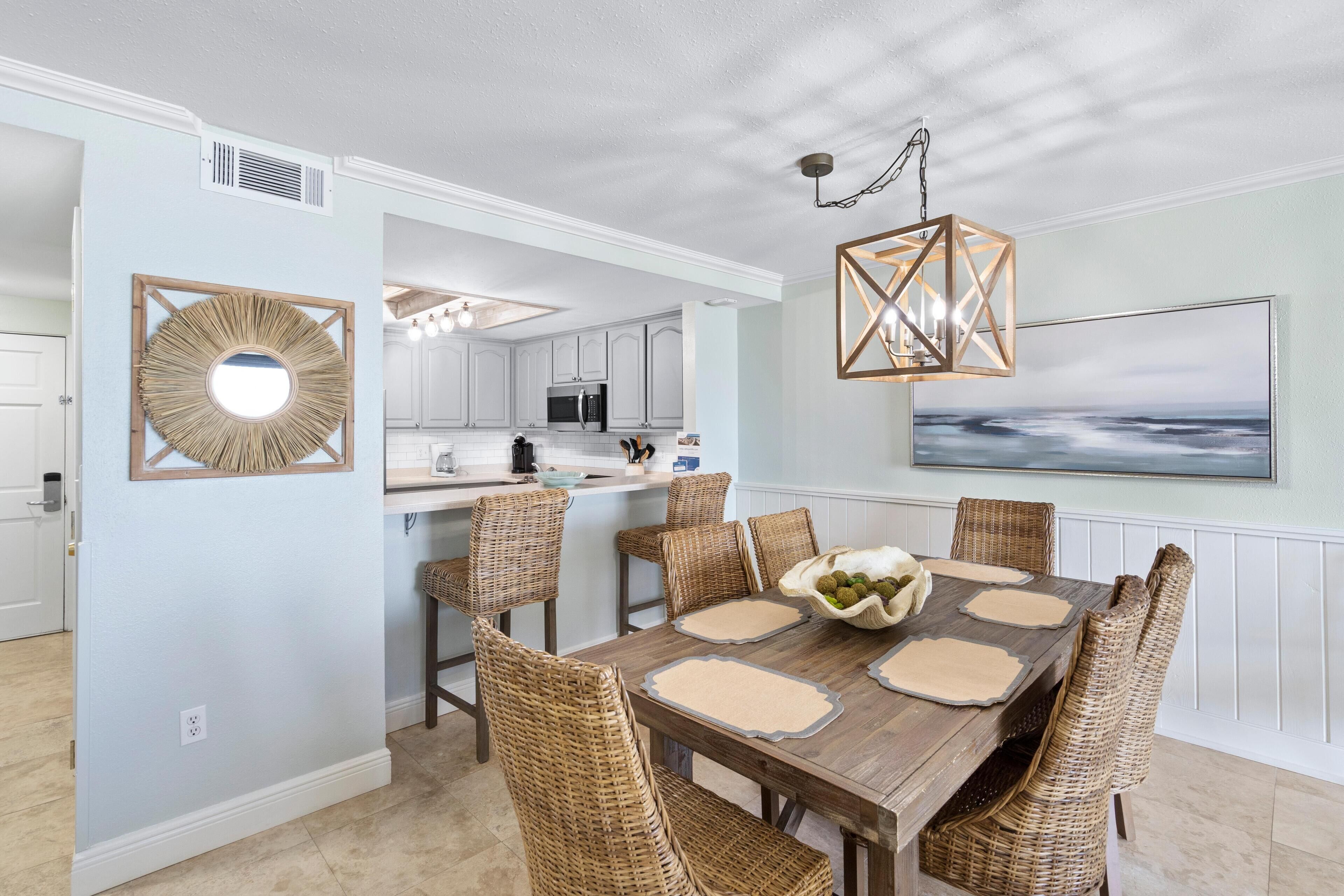 A dining area with a wooden table, wicker chairs, a central bowl of fruit, and a hanging light fixture.