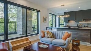 A modern living room with a grey sofa, wooden coffee table, and large windows offering a view of the outdoors.