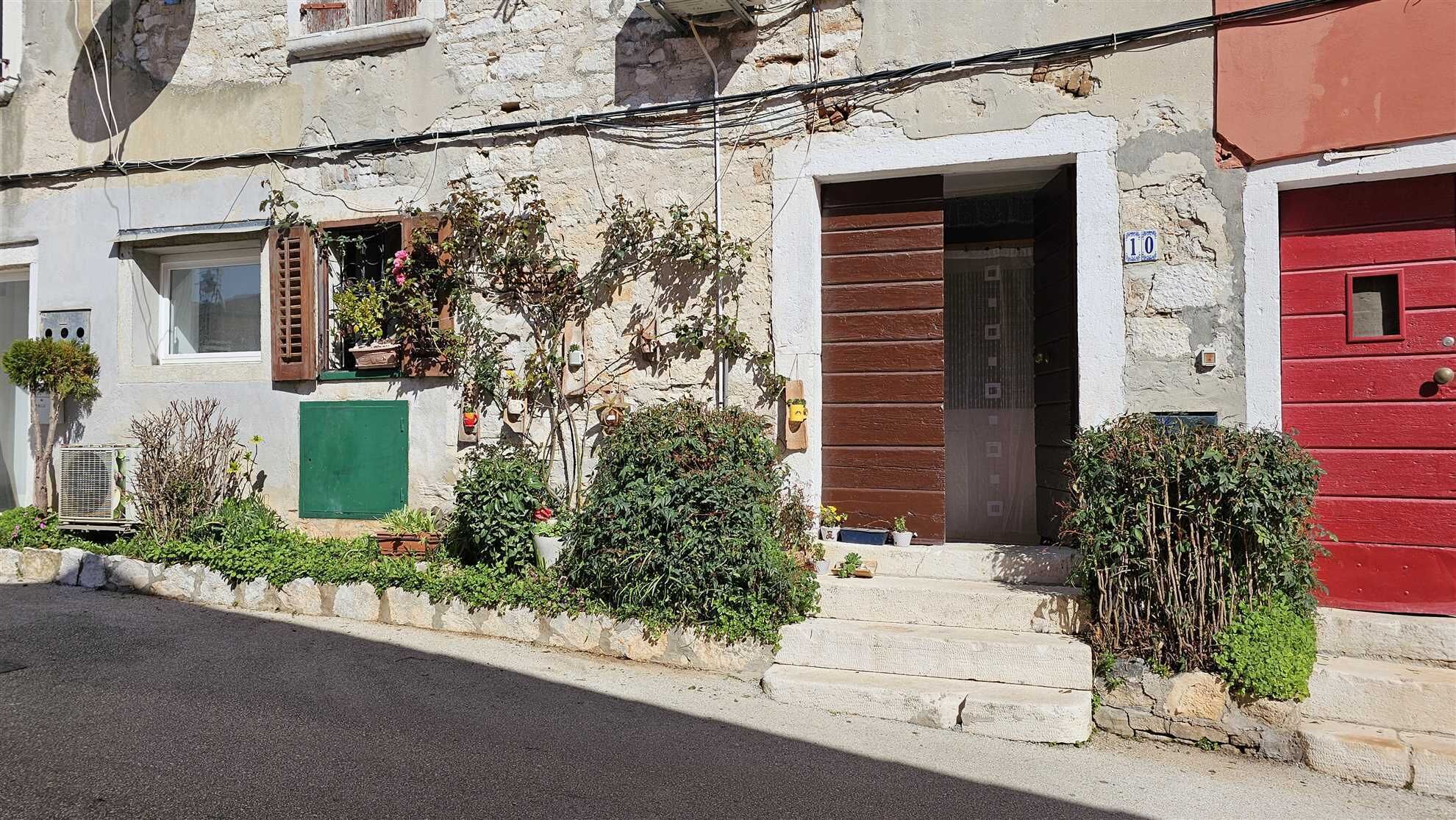 Un bâtiment à la façade en pierre, avec une porte en bois et une porte peinte en rouge. On y trouve des plantes et un petit jardin à l’avant.