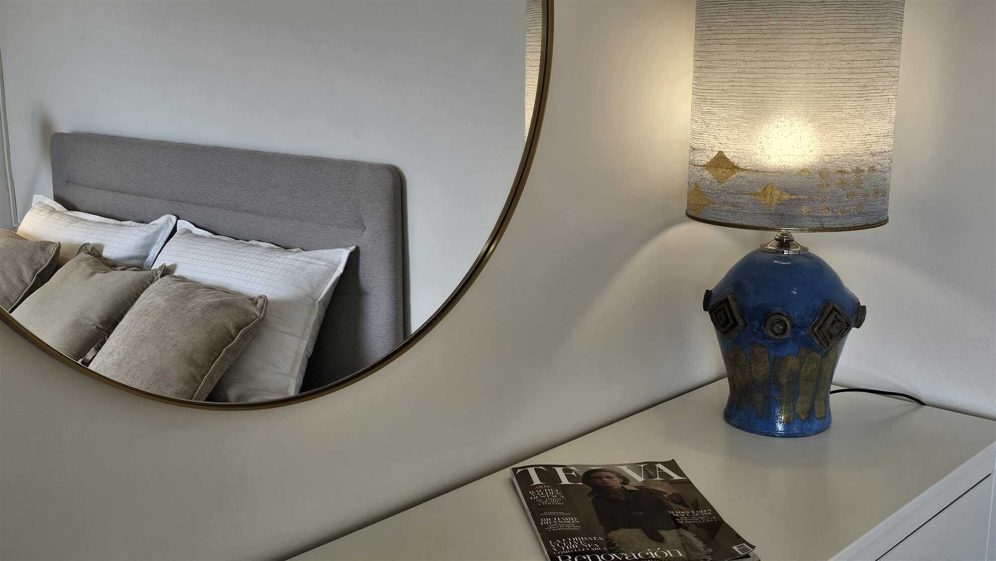 Un dormitorio con cama, almohadas, una mesita de noche, una lámpara y una revista.