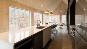 A modern kitchen with a large island, bar stools, and a view of the forest through large windows.