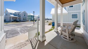 A covered porch with a wooden rocking chair, a white fence, and houses in the background. - Summer Salt 24181 (Orange Beach)