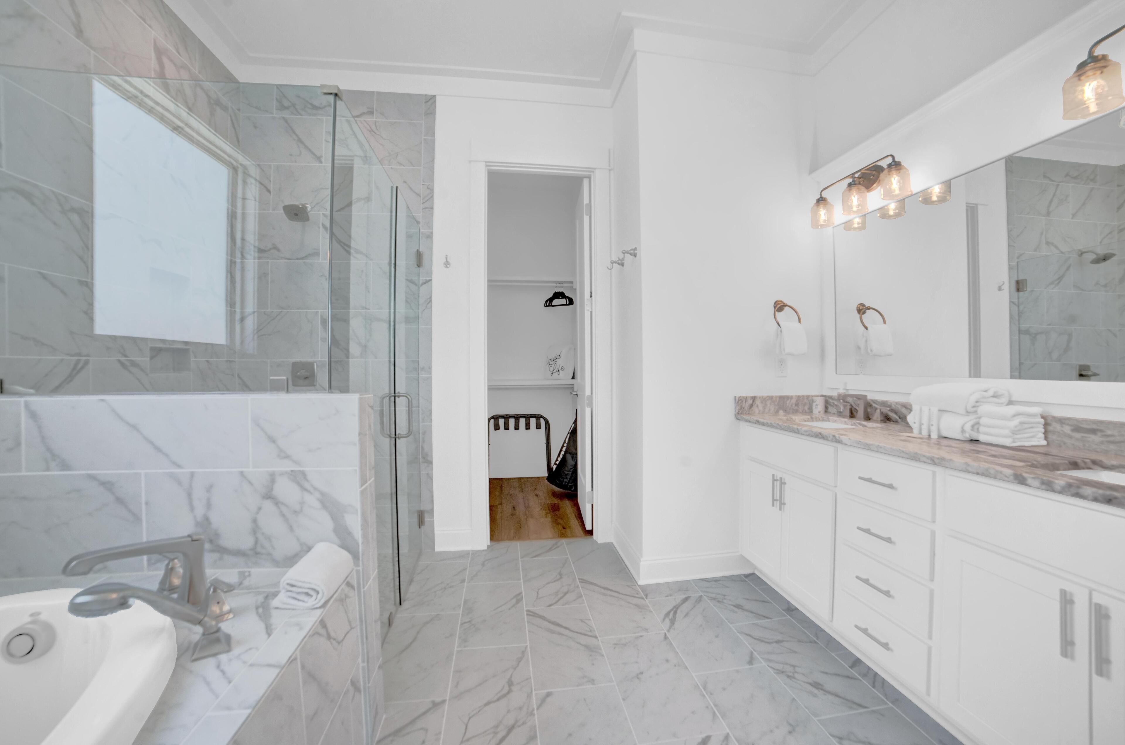 A modern bathroom with a glass-enclosed shower, a large mirror with lights, marble tiles, and white cabinetry.