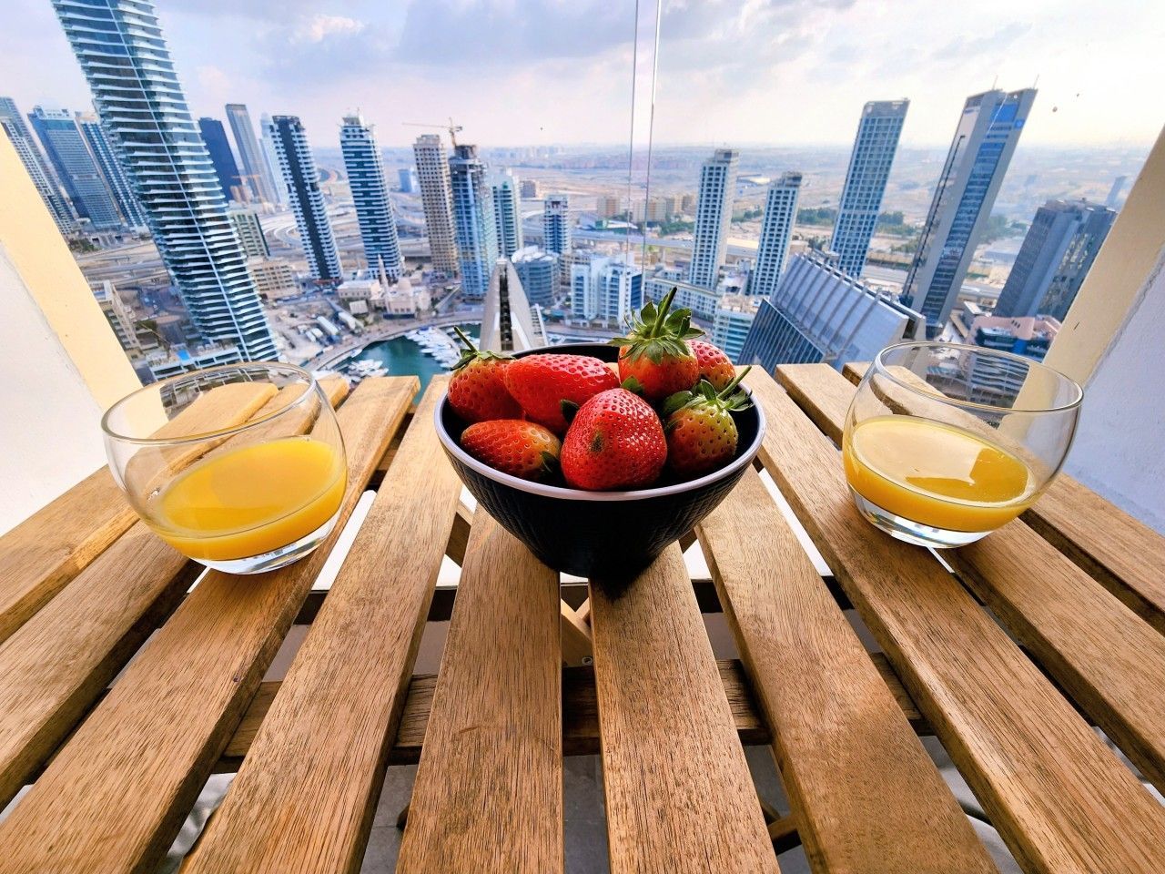 A wooden table with a bowl of strawberries and two glasses of orange juice, overlooking a cityscape with tall buildings.