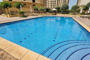 Una piscina rectangular con azulejos azules, rodeada de sillas de descanso y sombrillas, con edificios al fondo.