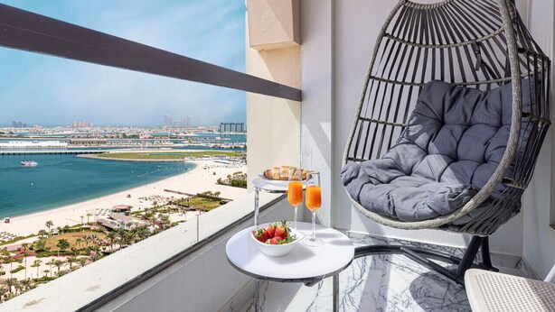 Een balkon met een hangstoel, een tafel met een fruitschaal en twee glazen sinaasappelsap, en uitzicht op een strand en stadsbeeld.