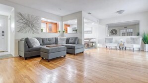 Ein geräumiges Wohnzimmer mit einem grauen Ecksofa, einem kleinen Hocker und einem Essbereich mit einem weißen Tisch und Stühlen.