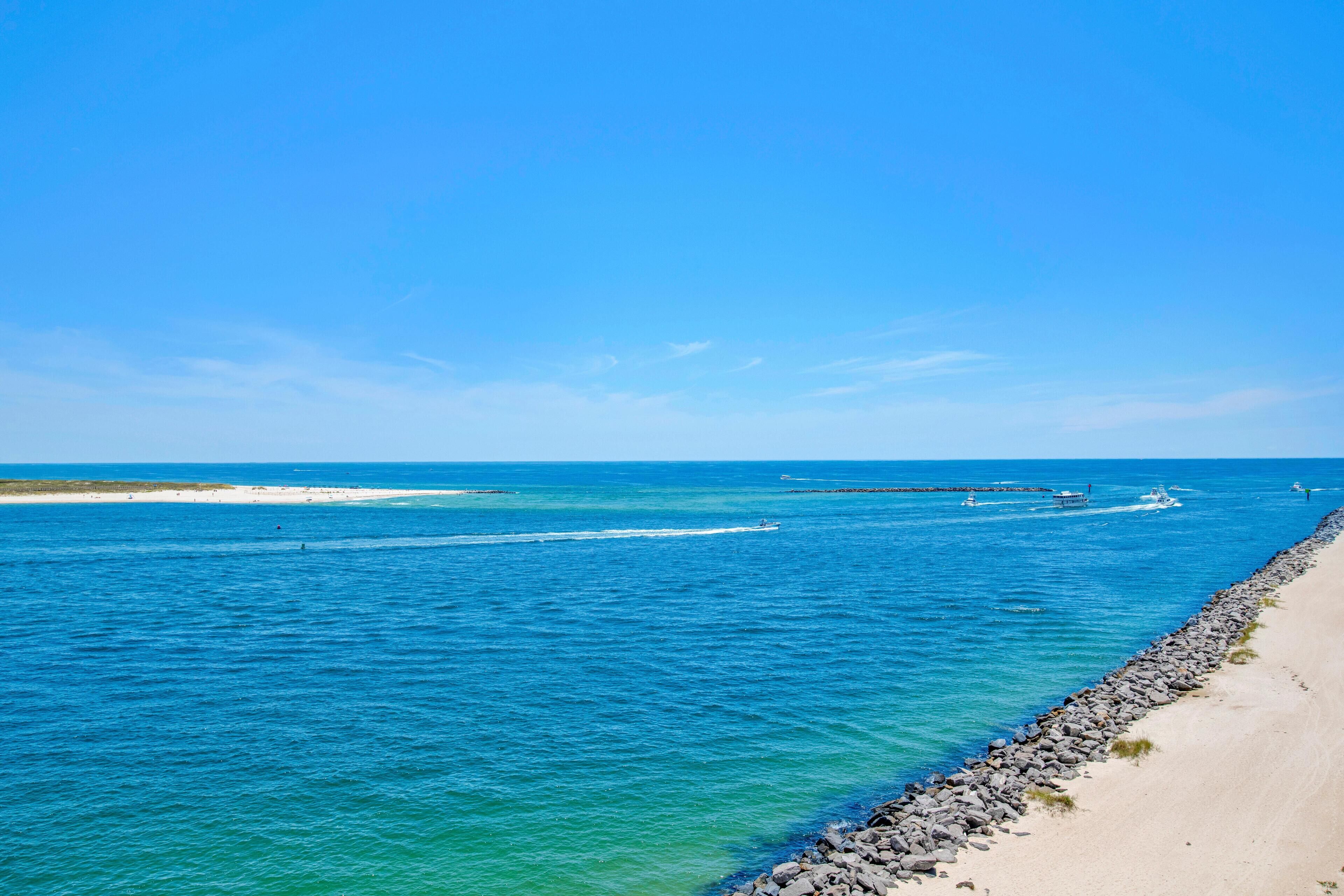 Escena costera con cielo azul despejado, mar tranquilo y playa arenosa con rompeolas rocosas.