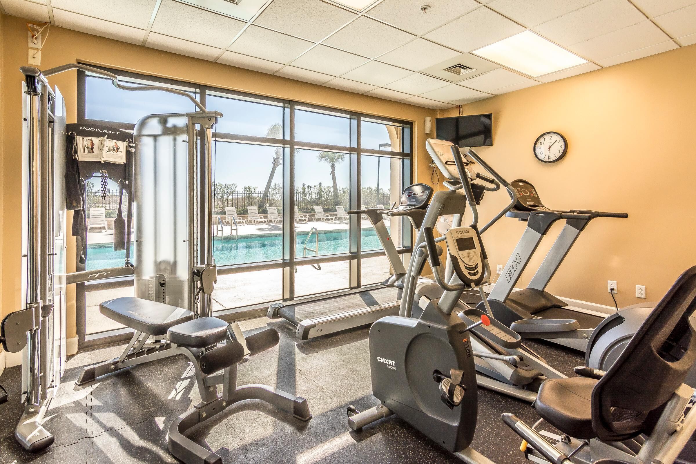 Un gimnasio con máquinas de cardio, equipo de pesas y un amplio ventanal con vistas a una piscina.