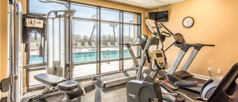 Un gimnasio con máquinas de cardio, equipo de pesas y un amplio ventanal con vistas a una piscina.