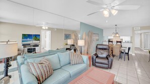 A spacious living area with a light blue sofa, a brown leather chair, and a dining table. A ceiling fan and a TV are visible in the background. - Gulf House 401 (Gulf Shores)