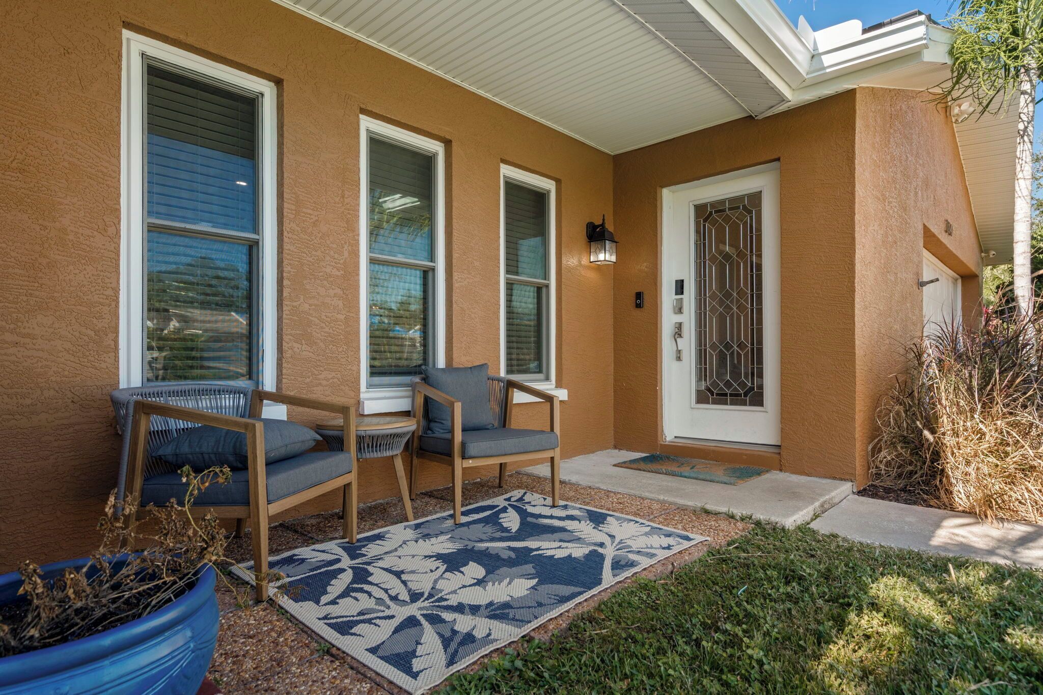 A house exterior with a porch, two chairs, a potted plant, and a patterned rug.