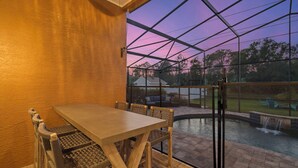 Un patio techado con mesa y sillas, vista a un jardín trasero con piscina y cascada, y un cielo al atardecer visible a través de la estructura.