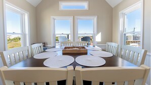 Uma sala de jantar com uma mesa grande, cadeiras brancas e vista para o exterior através de amplas janelas.