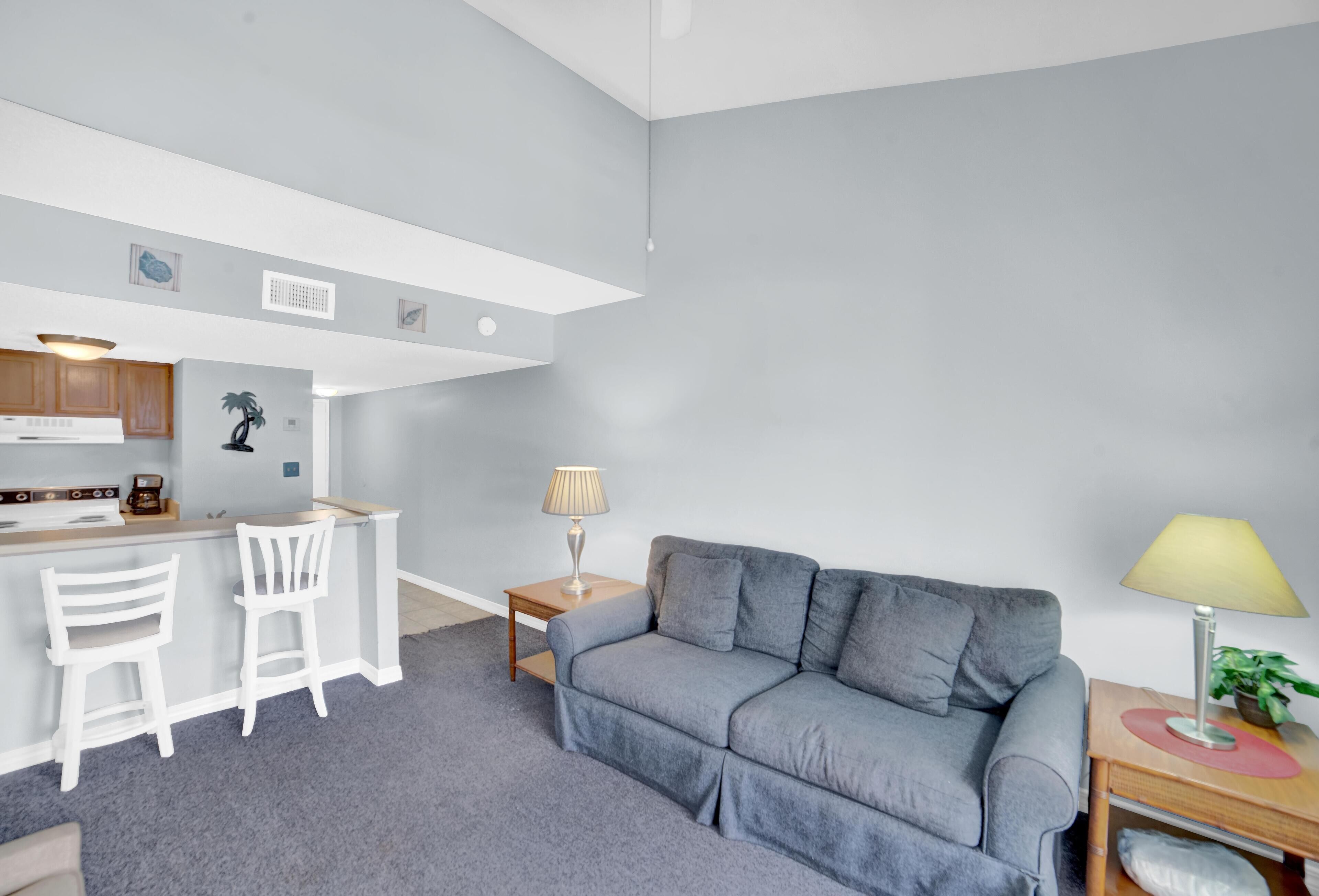 Een moderne woonkamer met een grijze bank, een klein houten tafeltje met een lamp, en een keuken met witte kasten en een bar.'