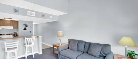 Een moderne woonkamer met een grijze bank, een klein houten tafeltje met een lamp, en een keuken met witte kasten en een bar.'