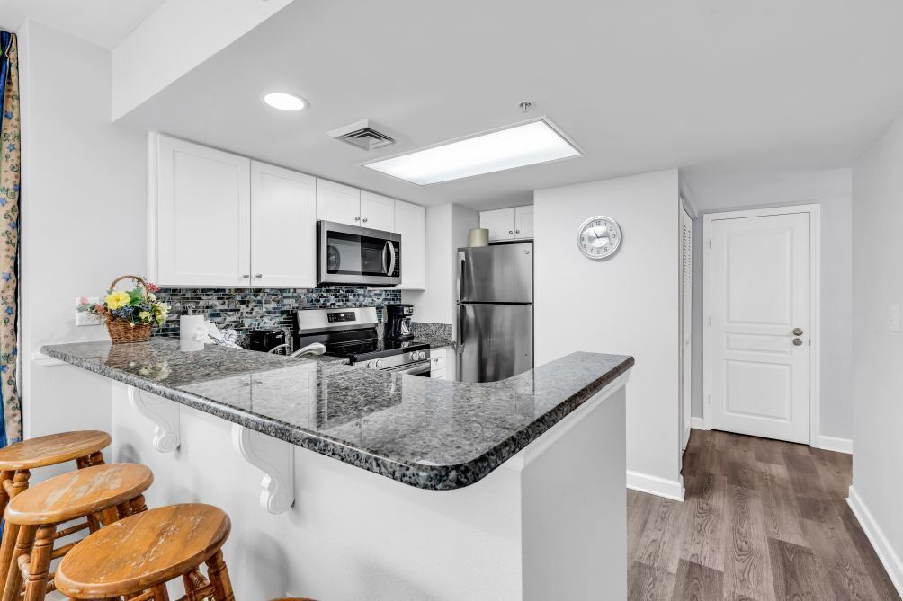 A modern kitchen with white cabinets, a granite countertop, a microwave, a refrigerator, and a clock on the wall.