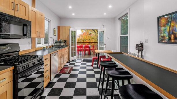 A modern kitchen with wooden cabinets, a black and white checkered floor, a dining area with red chairs, and a large window with a view of autumn trees.