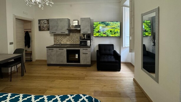 Cocina moderna con gabinetes grises, horno empotrado y campana extractora de acero inoxidable. Una silla negra está colocada junto a la cocina, y un televisor de pantalla plana grande está montado en la pared sobre la encimera.