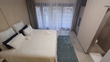 Quarto de hotel com cama, almofadas, toalha, tapete estampado e cortinas translúcidas.