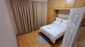 Een netjes opgemaakt bed met witte lakens en twee opgerolde handdoeken, een houten garderobe, een beige gordijn en een houten vloer.