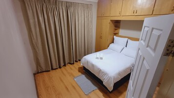 Uma cama bem arrumada com lençóis brancos e duas toalhas enroladas, um guarda-roupa de madeira, uma cortina bege e piso de madeira.