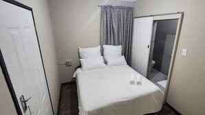 Een klein, net gemaakt bed met witte lakens in een compacte kamer met een deur en een raam met gordijnen.