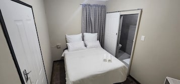 Cama pequena e bem arrumada com lençóis brancos em quarto compacto com porta e janela com cortinas.