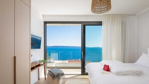 A modern bedroom with a large bed, a small seating area, and a view of the sea.