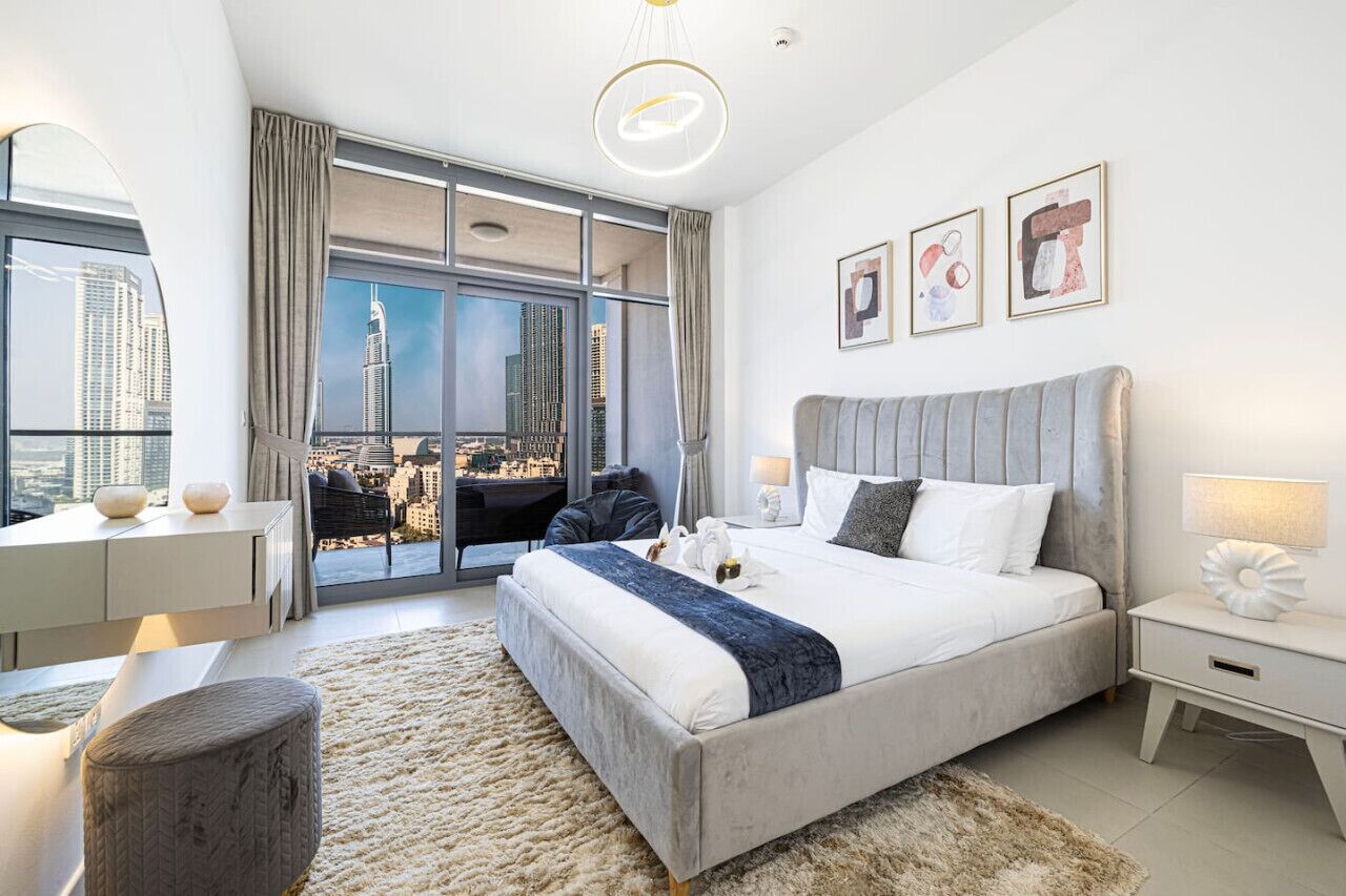 A modern hotel room with a large bed, a desk, a chair, and a view of a cityscape through floor-to-ceiling windows.