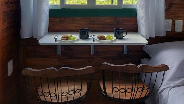 Ein Zimmer in einer Holzhütte mit Fensterblick auf eine grüne Landschaft, einem kleinen Tisch mit zwei Stühlen und einem Bett.