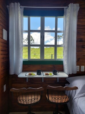Ein Zimmer in einer Holzhütte mit Fensterblick auf eine grüne Landschaft, einem kleinen Tisch mit zwei Stühlen und einem Bett.